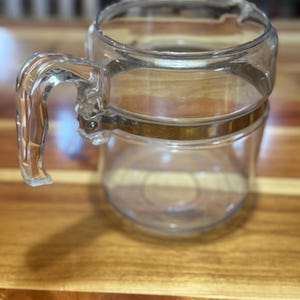 May include: A clear glass coffee pot with a handle and a gold-coloured band around the middle. The coffee pot is empty and sits on a wooden surface. The handle is attached to the side of the pot.
