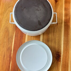 Puede incluir: Una olla de hierro fundido esmaltada en blanco con un interior de esmalte negro y una tapa separada. La olla y la tapa son redondas y tienen una superficie lisa.
