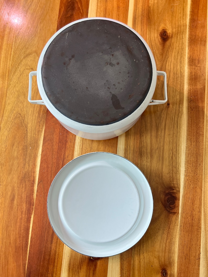Puede incluir: Una olla de hierro fundido esmaltada en blanco con un interior de esmalte negro y una tapa separada. La olla y la tapa son redondas y tienen una superficie lisa.