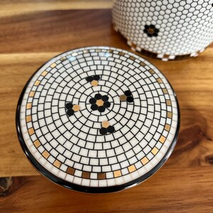 May include: A ceramic lid with a mosaic tile design featuring a central floral pattern in black and gold. The lid is surrounded by a black border. A matching ceramic cup with a honeycomb pattern is visible in the background.