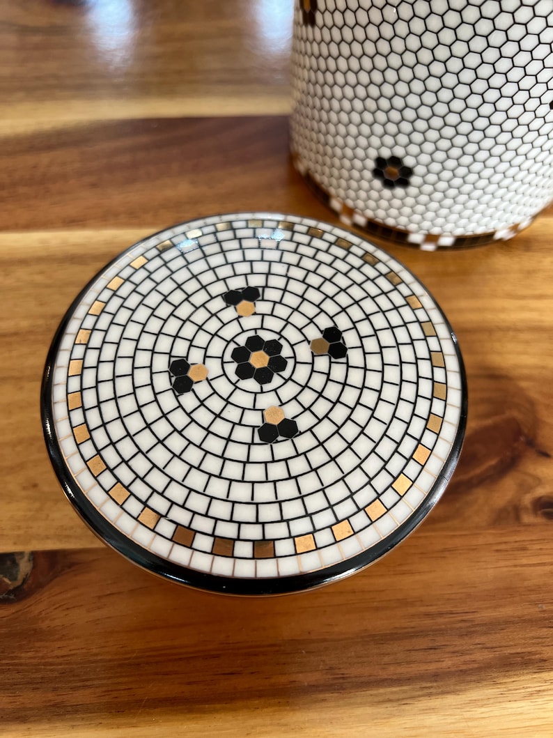 May include: A ceramic lid with a mosaic tile design featuring a central floral pattern in black and gold. The lid is surrounded by a black border. A matching ceramic cup with a honeycomb pattern is visible in the background.