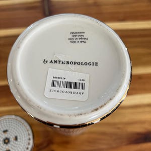 May include: A white ceramic Anthropologie container with a gold rim and a black and white checkered pattern. The bottom of the container has a label with the brand name and a barcode. The lid is visible in the lower left corner.