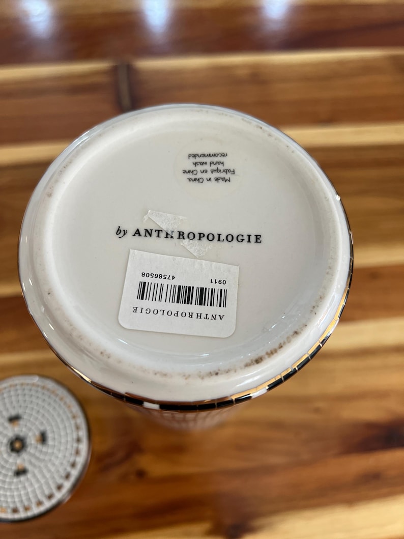 May include: A white ceramic Anthropologie container with a gold rim and a black and white checkered pattern. The bottom of the container has a label with the brand name and a barcode. The lid is visible in the lower left corner.