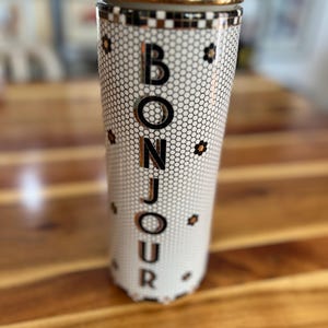 May include: A tall, cylindrical container with a gold-colored lid and rim. The container is white with a black honeycomb pattern and the word "BONJOUR" in large, gold-colored letters. Small black and gold flower accents are scattered around the container.