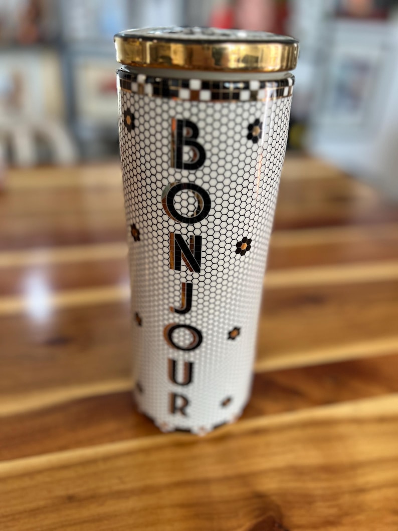 May include: A tall, cylindrical container with a gold-colored lid and rim. The container is white with a black honeycomb pattern and the word "BONJOUR" in large, gold-colored letters. Small black and gold flower accents are scattered around the container.