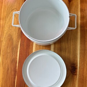 Puede incluir: Olla de esmalte blanco con dos asas y una tapa a juego. La olla y la tapa tienen un borde negro.