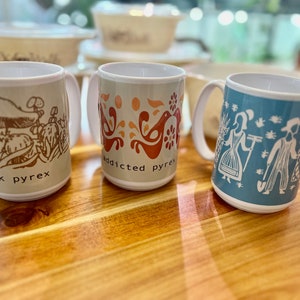 May include: Three ceramic mugs with different designs. The first mug is beige with a brown mushroom design and the text "peak pyrex". The second mug is beige with a red and orange bird design and the text "addicted pyrex". The third mug is blue with a white design of two people gardening and the text "pyrex".