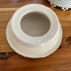 May include: Cream-colored ceramic lid with a gold rim. The lid has a circular opening in the center and a slightly raised edge. The lid is likely for a jar or container, and is sitting on a wooden surface.