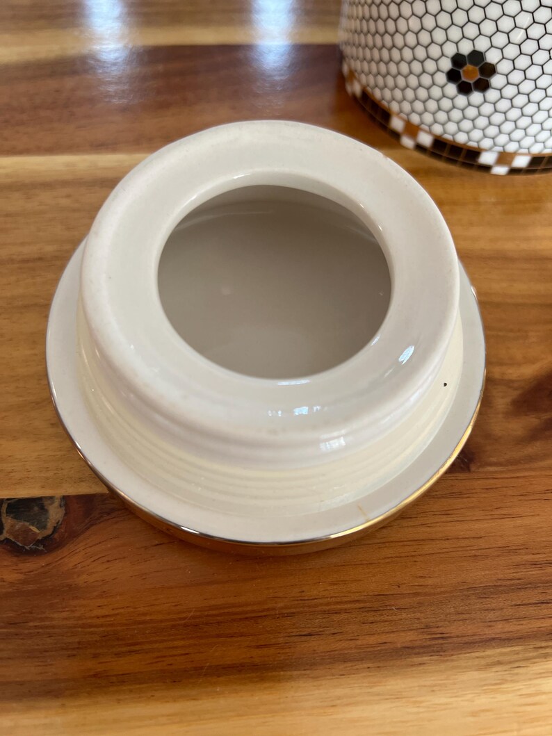 May include: Cream-colored ceramic lid with a gold rim. The lid has a circular opening in the center and a slightly raised edge. The lid is likely for a jar or container, and is sitting on a wooden surface.