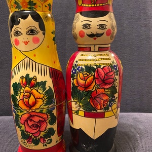 May include: Two traditional Russian nesting dolls, one depicting a woman in a yellow headscarf and the other a man in a red hat and military uniform. Both dolls are painted with floral designs and the woman's doll has the word "Rodnik" written on the base.