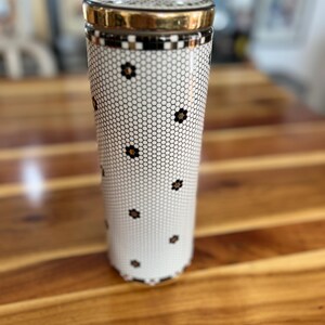 May include: A tall, cylindrical container with a gold-colored lid and rim. The body features a white background with a black honeycomb pattern and small black flower accents. The container sits on a wooden surface.
