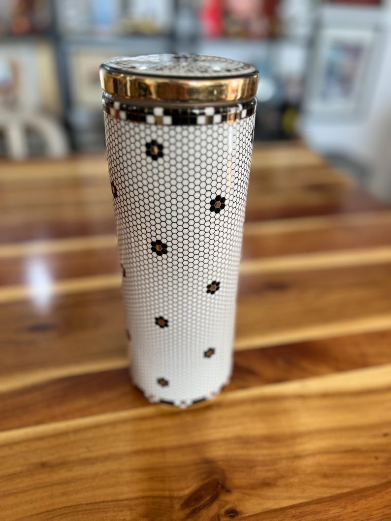 May include: A tall, cylindrical container with a gold-colored lid and rim. The body features a white background with a black honeycomb pattern and small black flower accents. The container sits on a wooden surface.