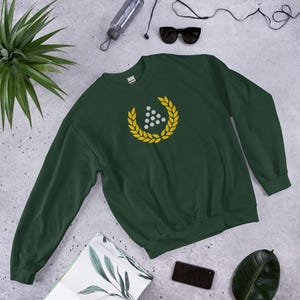 Può includere: Una felpa a maniche lunghe color verde bosco scuro con una grafica centrale raffigurante una corona d'alloro gialla che circonda un gruppo di sei punti bianchi disposti a triangolo sul petto del capo.