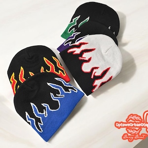 Puede incluir: Cinco gorros de punto con dise&ntilde;os de llamas en varios colores, incluyendo negro, blanco, azul, rojo y verde. Los gorros est&aacute;n dispuestos sobre una superficie blanca.