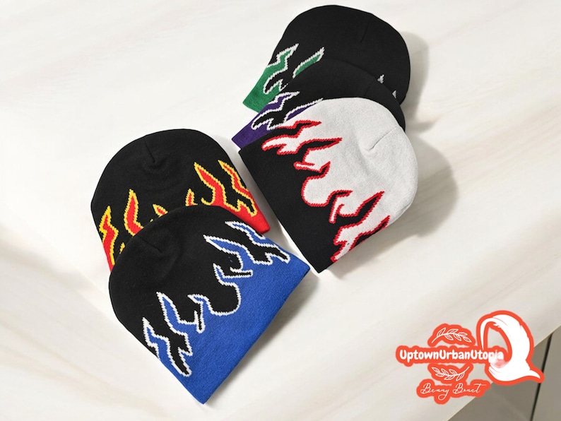 Puede incluir: Cinco gorros de punto con dise&ntilde;os de llamas en varios colores, incluyendo negro, blanco, azul, rojo y verde. Los gorros est&aacute;n dispuestos sobre una superficie blanca.