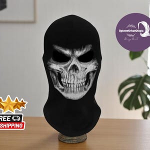 Könnte beinhalten: Schwarze Sturmhaube mit einem weißen Totenkopf-Design. Der Schädel hat einen bedrohlichen Ausdruck mit einem breiten Grinsen und scharfen Zähnen.