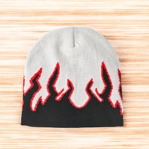 Puede incluir: Un gorro gris con un dise&ntilde;o de llamas negras y rojas. El gorro est&aacute; hecho de un tejido suave y de punto.