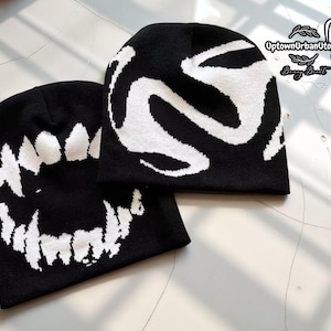 Può includere: Due berretti neri a maglia con disegni grafici bianchi. Un berretto presenta un sorriso bianco e dentato, mentre l'altro ha un motivo astratto bianco. I berretti sono esposti su una superficie di colore chiaro.