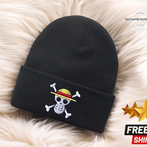 Puede incluir: Gorro negro con una calavera blanca y huesos cruzados bordados en la parte delantera. La calavera lleva un sombrero de paja amarillo y rojo. El gorro está sobre una superficie blanca y esponjosa.