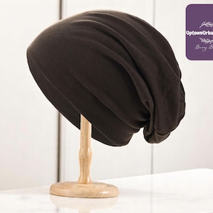 Puede incluir: Un gorro holgado de color marrón oscuro exhibido en un soporte de madera. El gorro tiene un estilo relajado y holgado y parece estar hecho de una tela suave. El logotipo de UptownUrbanUtopia es visible en la esquina superior derecha.