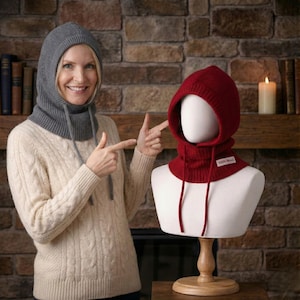 May include: A woman points to a gray knit hood and a red knit hood on a mannequin. The hoods have drawstrings and are made of a thick, warm material. The red hood has a small white tag that reads "100% Wool".