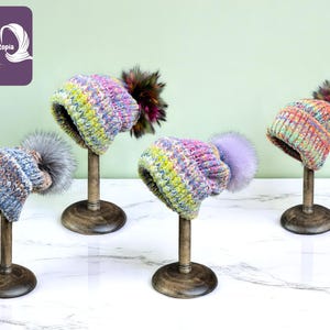 Puede incluir: Cuatro gorros de punto coloridos con pompones de piel sintética se exhiben en soportes de madera. Los gorros presentan una mezcla de colores, incluyendo azul, rosa, amarillo y naranja. Los soportes están sobre una superficie de mármol blanco, con un fondo verde.