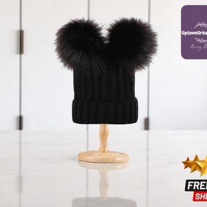 Può includere: Un berretto nero a maglia con due grandi e soffici pompon di pelliccia nera. Il cappello è esposto su un supporto di legno. Un accessorio invernale su sfondo bianco.