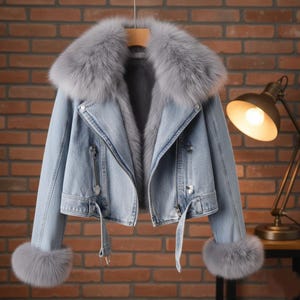 Puede incluir: Chaqueta vaquera azul claro con cuello y puños de piel gris. La chaqueta tiene cierre de cremallera, trabillas y cinturón. La chaqueta está colgada de una percha de madera.