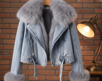 Peut inclure: Veste en jean bleu clair avec un col et des poignets en fourrure grise. La veste a une fermeture éclair, des passants de ceinture et une ceinture. La veste est suspendue à un cintre en bois.