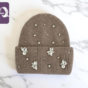May include: A brown knit beanie adorned with sparkling rhinestones. The hat features a folded brim and is embellished with various sizes of clear, round rhinestones and floral-shaped crystal accents. The hat is displayed on a white marble surface.