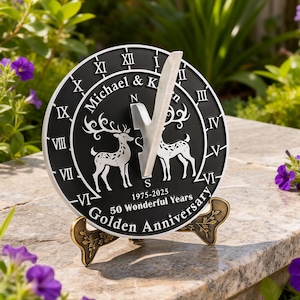 Könnte beinhalten: Eine schwarze und silberne Sonnenuhr mit römischen Ziffern und dem Text "Michael & K" und "50 Wonderful Years Golden Anniversary". Auf dem Zifferblatt sind zwei Hirsche abgebildet. Die Sonnenuhr wird von einem dekorativen Ständer getragen.