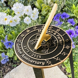 May include: A black and gold sundial with Roman numerals and the text "Megan & Parker" and "Golden Anniversary 1975-2025". The sundial features a gold gnomon and decorative bells and doves. Surrounded by white and purple flowers.
