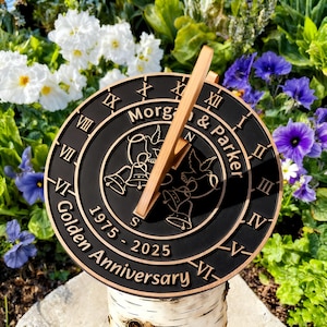 May include: A black and bronze sundial with Roman numerals and the text "Morgan & Parker Golden Anniversary 1975-2025." The sundial features a wooden gnomon and decorative doves and bells. It is mounted on a birch log.