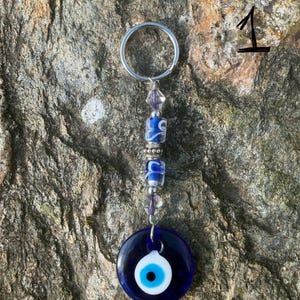 May include: A blue and white evil eye keychain with a silver keyring. The keychain features a dark blue glass eye charm with a white and light blue eye design. The charm is connected to a beaded strand with blue and clear beads.