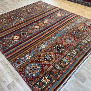 Könnte beinhalten: Ein rechteckiger Teppich mit geometrischem Muster. Der Teppich hat einen roten Hintergrund mit Streifen in Blau, Orange und Grün. Auf der Oberfläche sind komplizierte Blumen- und geometrische Muster eingearbeitet. Der Teppich hat einen Fransenrand.