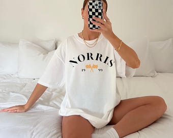 May include: White oversized t-shirt with black text that reads "NORRIS" in an arc with checkered flags and the numbers "19" and "99" underneath.