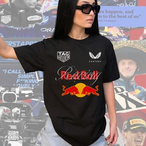 Könnte beinhalten: Schwarzes T-Shirt mit einem weißen Red Bull Logo und dem Text "TAG HEUER" und "CASTORE" auf der Vorderseite. Das Shirt zeigt außerdem zwei rote Bullen mit gelben Augen.