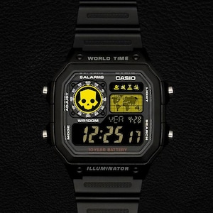 May include: A black digital Casio watch with a resin strap, featuring a square face with a yellow skull graphic, a world map display, and the text World Time, 5 Alarms, WR100M, 10 Year Battery, and Illuminator printed on the watch case.