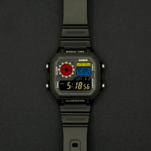 May include: A black Casio digital watch with a rectangular face and a black band. The watch face displays the time, date, and world time features. The watch has a red and blue circular design on the face.