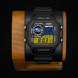 May include: A black Casio digital watch with a square face, featuring a Subaru WRX logo, world time map, and a yellow LCD display showing 8:03:17. The watch includes text labels for 5 alarms, WR100M, 10 year battery, and illuminator on the bezel.