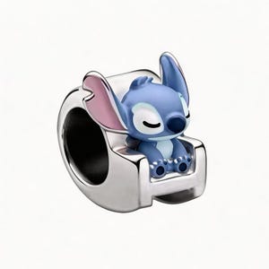May include: A silver charm featuring a blue cartoon character with large ears and a small nose, seated in a silver chair. The character has closed eyes and pink accents on the ears. The charm is a collectible item.