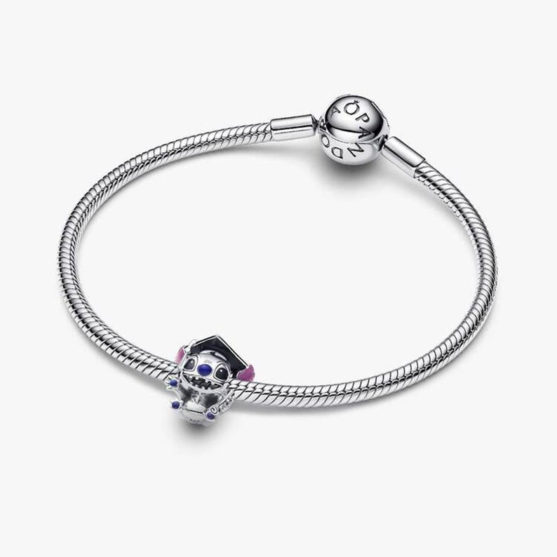 May include: A silver snake chain bracelet featuring a round clasp engraved with the word PANDORA. A charm shaped like a blue and purple character wearing a black graduation cap is threaded onto the bracelet, adding a distinct decorative element.