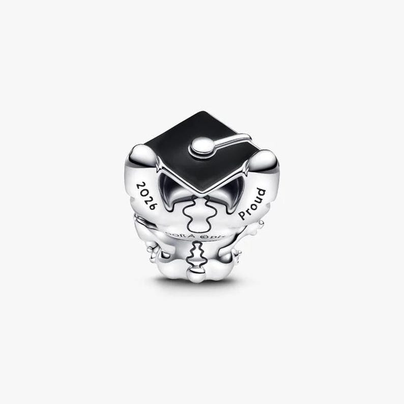 May include: A silver charm bead shaped like a character wearing a black enamel graduation cap. The charm features the engraved text 2026 on one side and the word Proud on the other side, with a central vertical puzzle piece design detail.