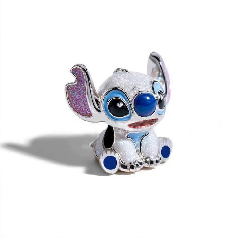 May include: A small, glittery white charm shaped like the character Stitch, featuring large ears with pink glitter interiors, a dark blue nose, and black eyes. The figure has blue accents on the feet and chest, with a metallic silver finish.