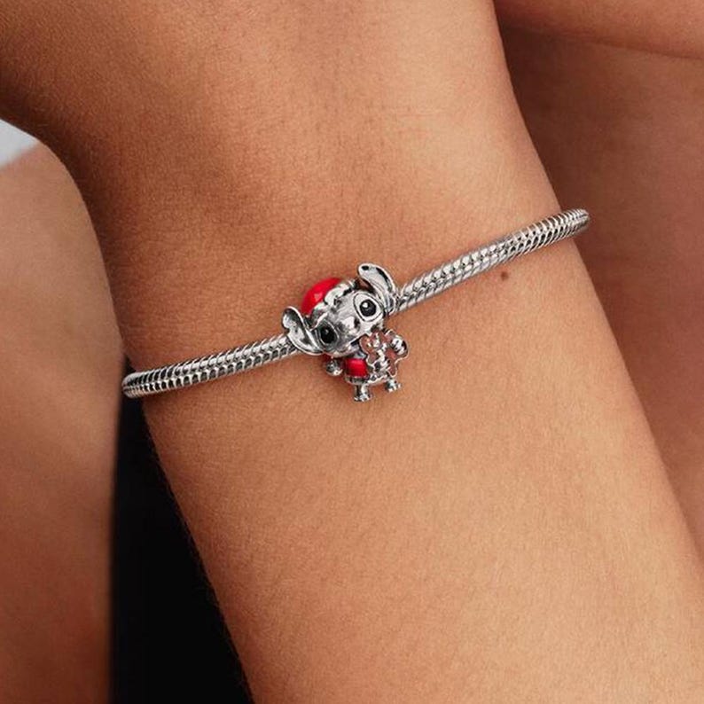 Op de afbeelding: Een zilveren bedelarmband met een gedetailleerde bedel van een stripfiguur met een rode kerstmuts en rode accenten. De armband heeft een slangenkettingontwerp en wordt om een pols gedragen.