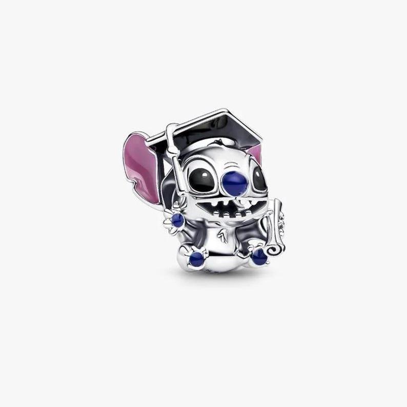 May include: A silver charm shaped like the character Stitch wearing a black graduation cap. The charm features pink enamel on the inner ears, a dark blue nose, and small blue stones set into the hands and feet. Stitch is holding a diploma.