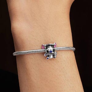 May include: A silver charm bracelet featuring a character charm of a creature wearing a graduation cap. The charm has pink inner ears, black eyes, and small blue accents on the hands and nose, attached to a textured, flexible silver chain.