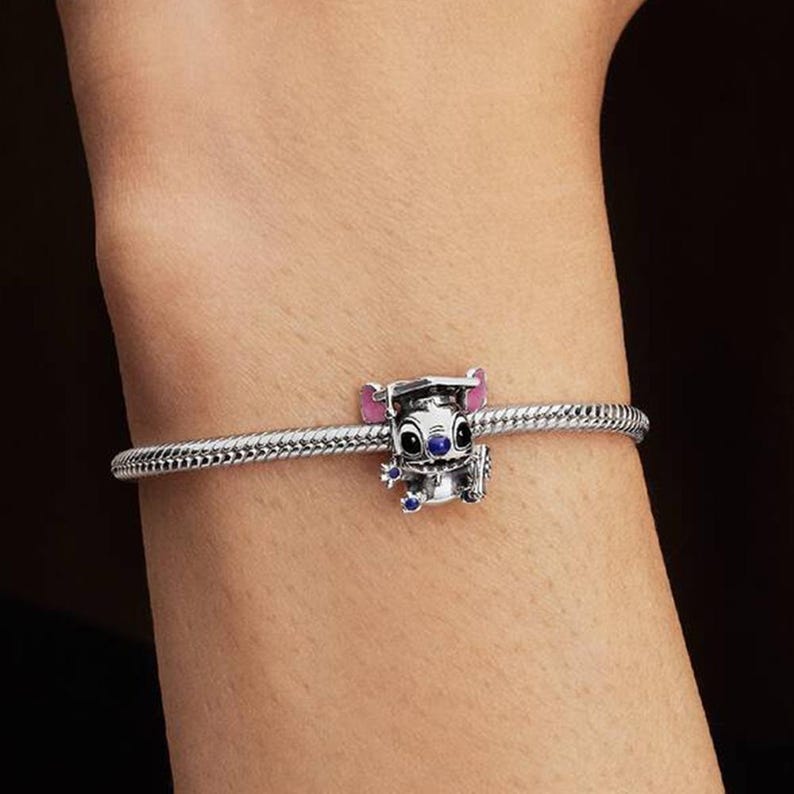 May include: A silver charm bracelet featuring a character charm of a creature wearing a graduation cap. The charm has pink inner ears, black eyes, and small blue accents on the hands and nose, attached to a textured, flexible silver chain.