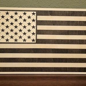 May include: A wooden American flag with a dark brown stain. The stars are cut out of the wood, creating a negative space effect.