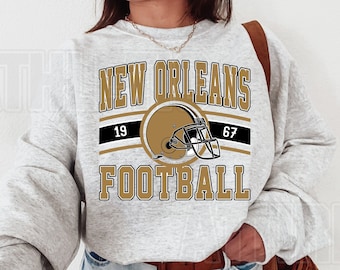 May include: A gray sweatshirt with the text "NEW ORLEANS FOOTBALL" in gold lettering. A black and gold football helmet graphic is centered above the text. The numbers "19" and "67" are on either side of the helmet.